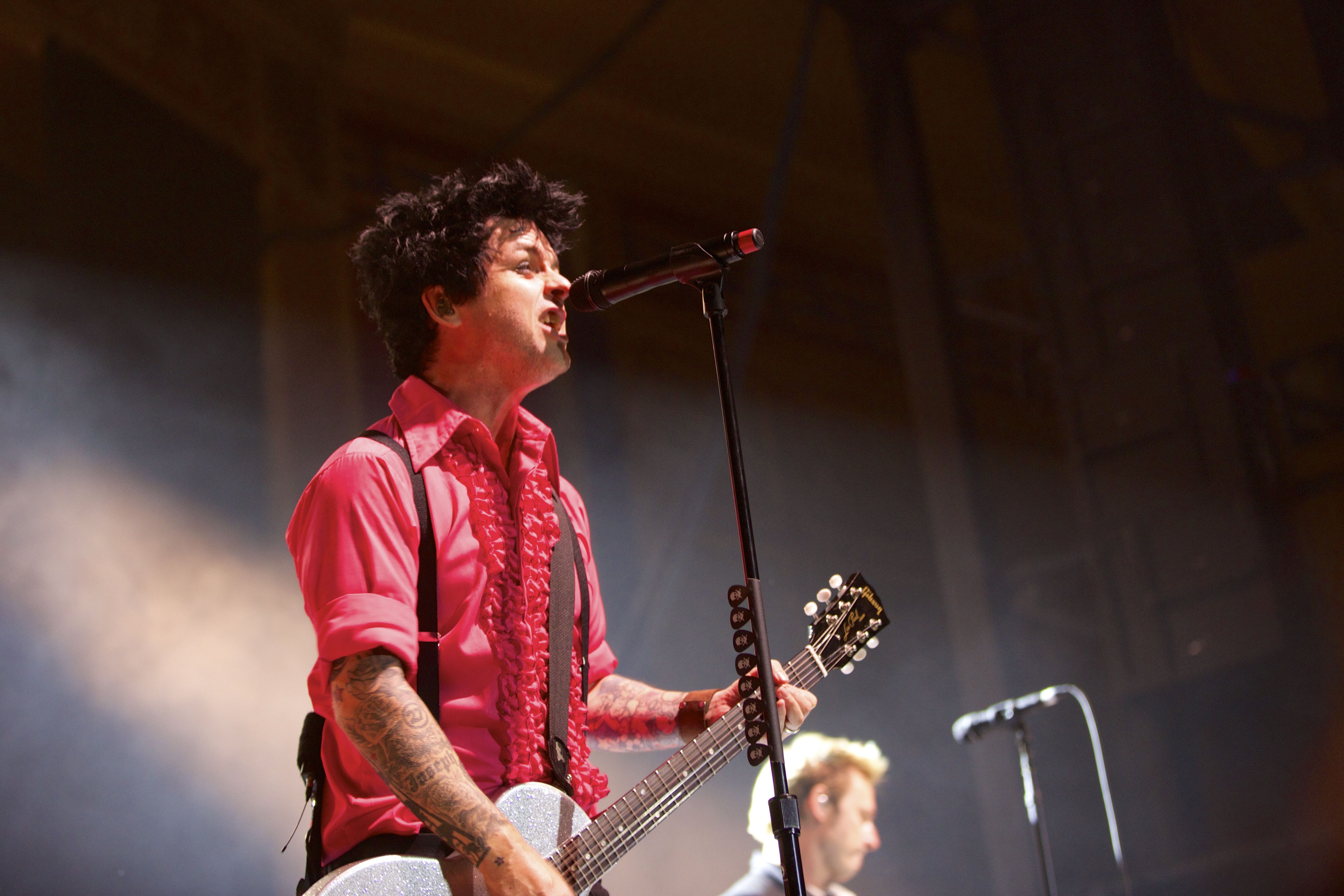 A white man with black hair sings on stage, while playing a white guitar and wearing a red shirt and black suspenders.