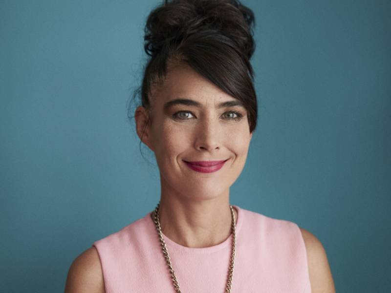 A white woman with dark brown hair held up in a messy up-do, stands in front of a blue-green wall.