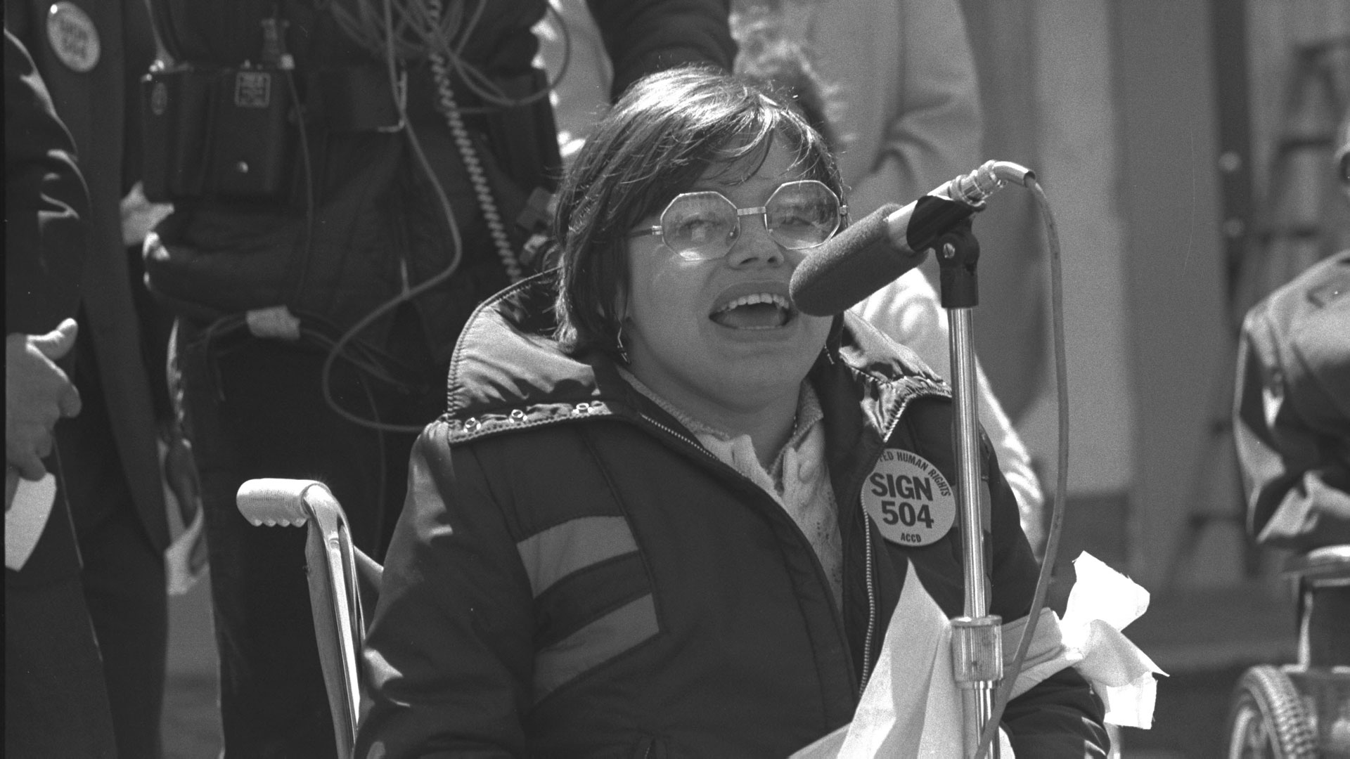 A woman in a wheelchair speaks enthusiastically into a microphone. She is wearing an anorak and glasses.