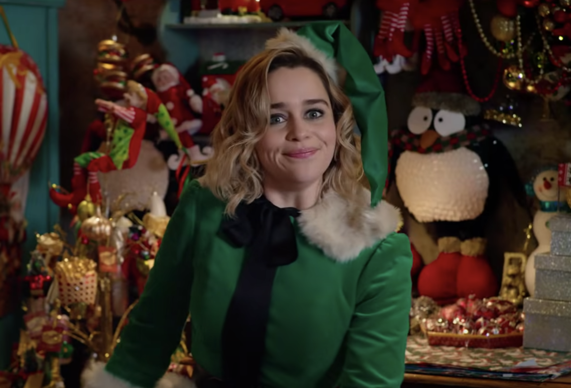 A young woman dressed in a green elf suit smiles in a room filled with festive ornaments and decorations.