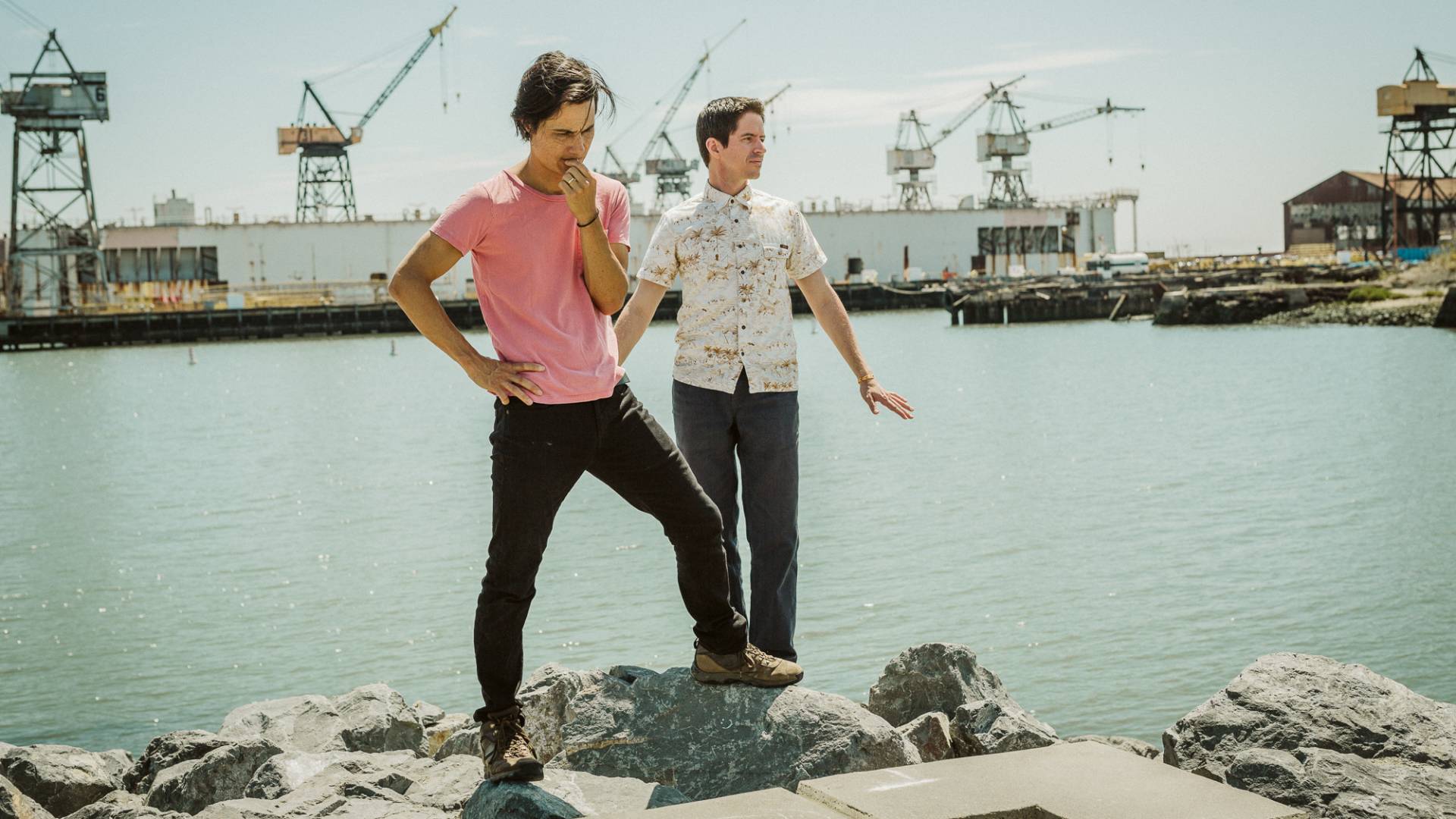 Two men stand on a shoreline, industrial cranes lines up across the water from them.