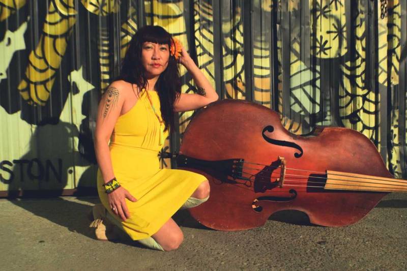 Jazz musician Caroline Chung wear a bright yellow dress as she bends down, posing next to her upright bass.