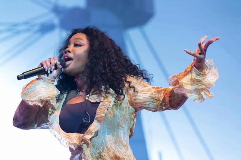 A Black woman with long wavy hair sings into a microphone on stage and gestures.