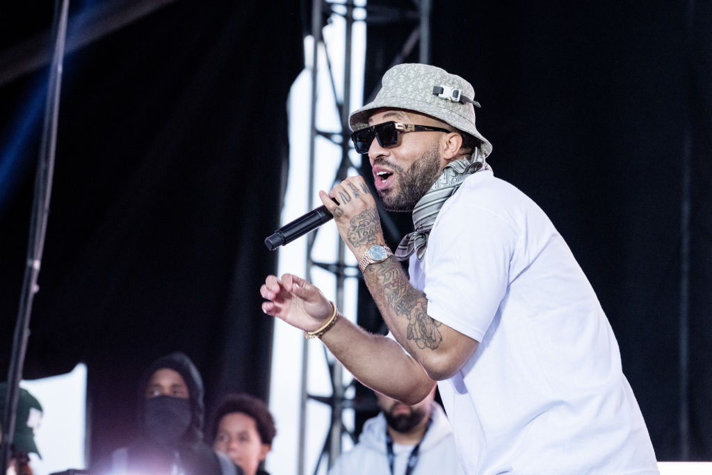 Larry June raps into the microphone on a big festival stage. He's wearing a bucket hat, designer sunglasses and a bandana and is smiling.