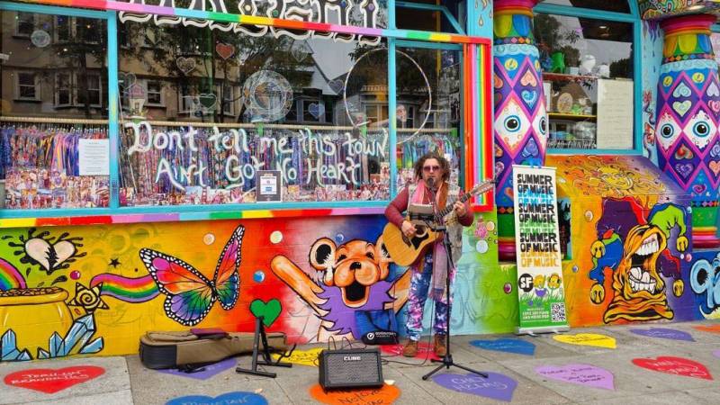 Guitar player sings into mic in front of brightly painted psychedelic storefront.