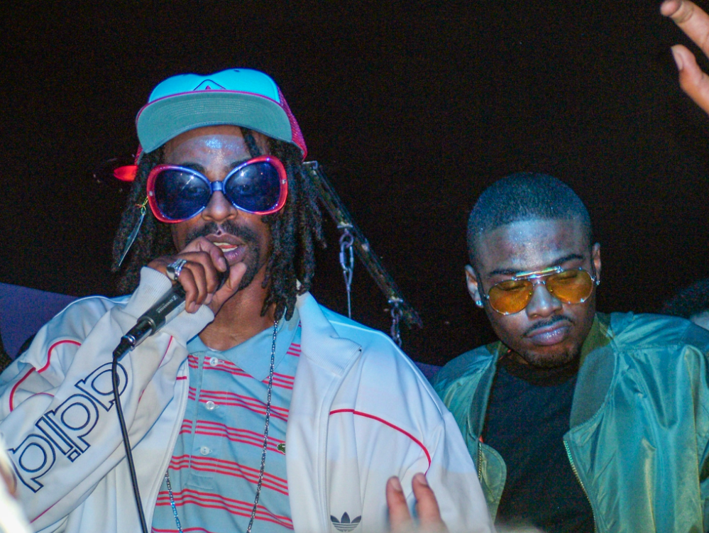 Mac Dre performs on stage in the early 2000s, wearing giant sunglasses, a striped polo shirt and Adidas jacket.