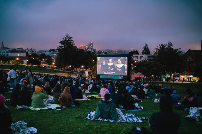 Nighttime view of crowd on blankets in Dolores Park, large inflated screen in distance with three nuns and dialogue below.