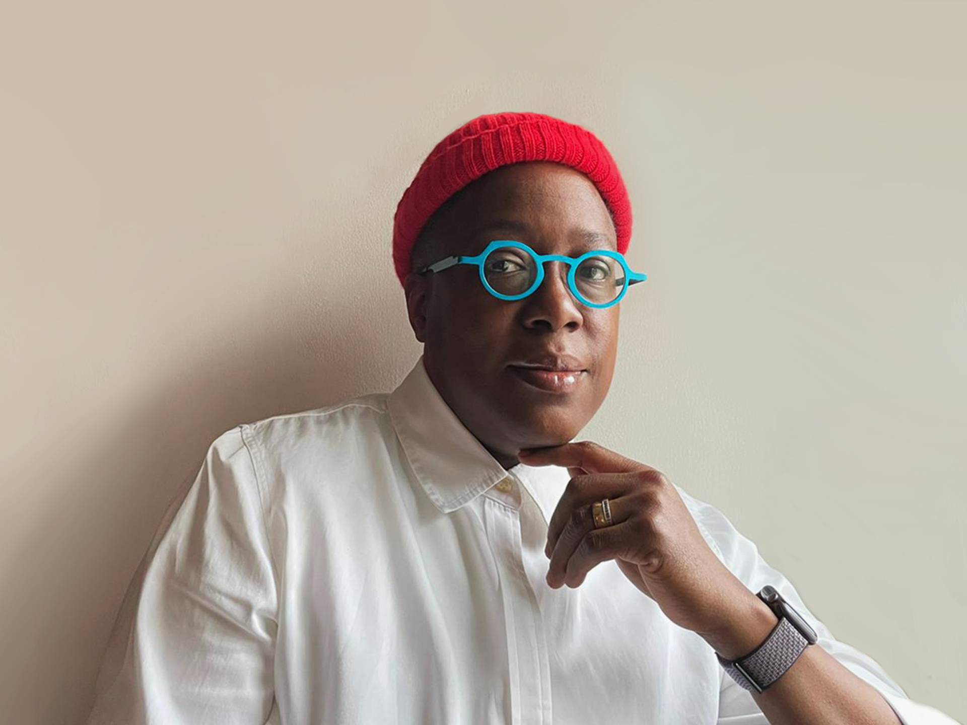 A Black woman sits against a white wall wearing a white buttoned up shirt, small, round, green eye glasses and a red beanie. She rests one finger under her chin.