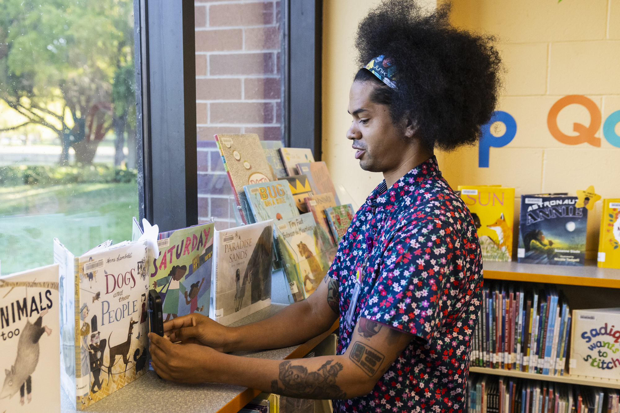 A person with their hair in an afro leans a cell phone up against sone books on a shelf in an indoor setting.