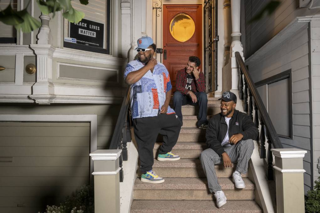 Three people sit and lean on the steps outside of a home and smile.