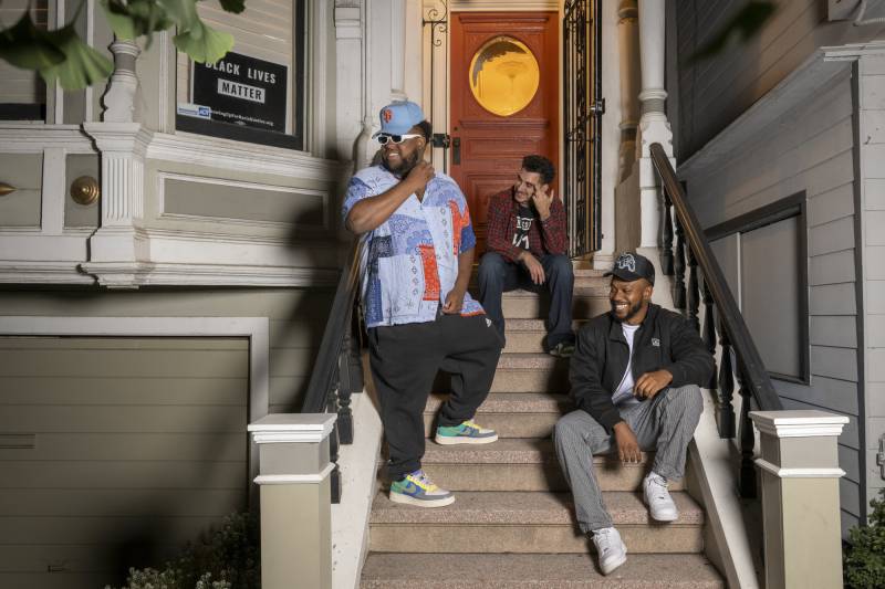 Three people sit and lean on the steps outside of a home and smile.