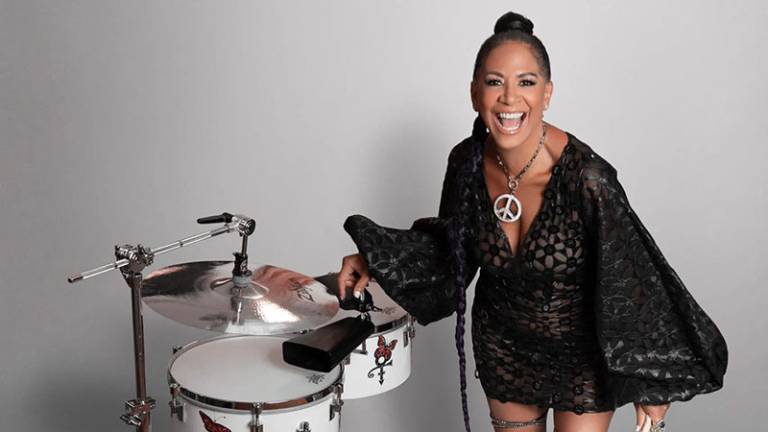 A woman in black smiles into the camera near a set of timbales