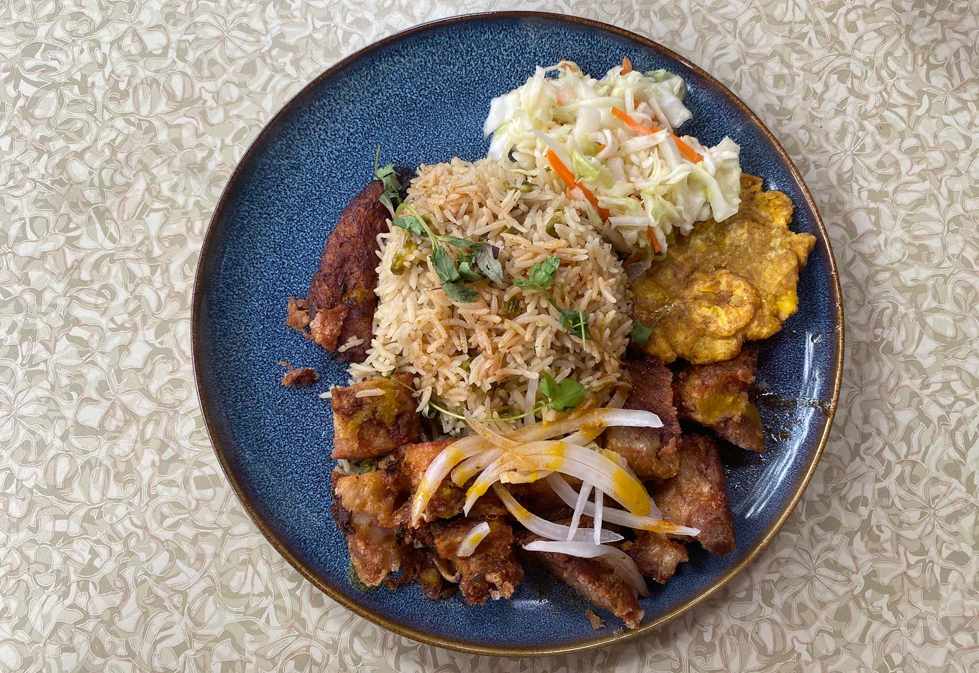 Fried pork, rice, plantains and pikliz (pickled cabbage and carrots) on a dark blue plate.