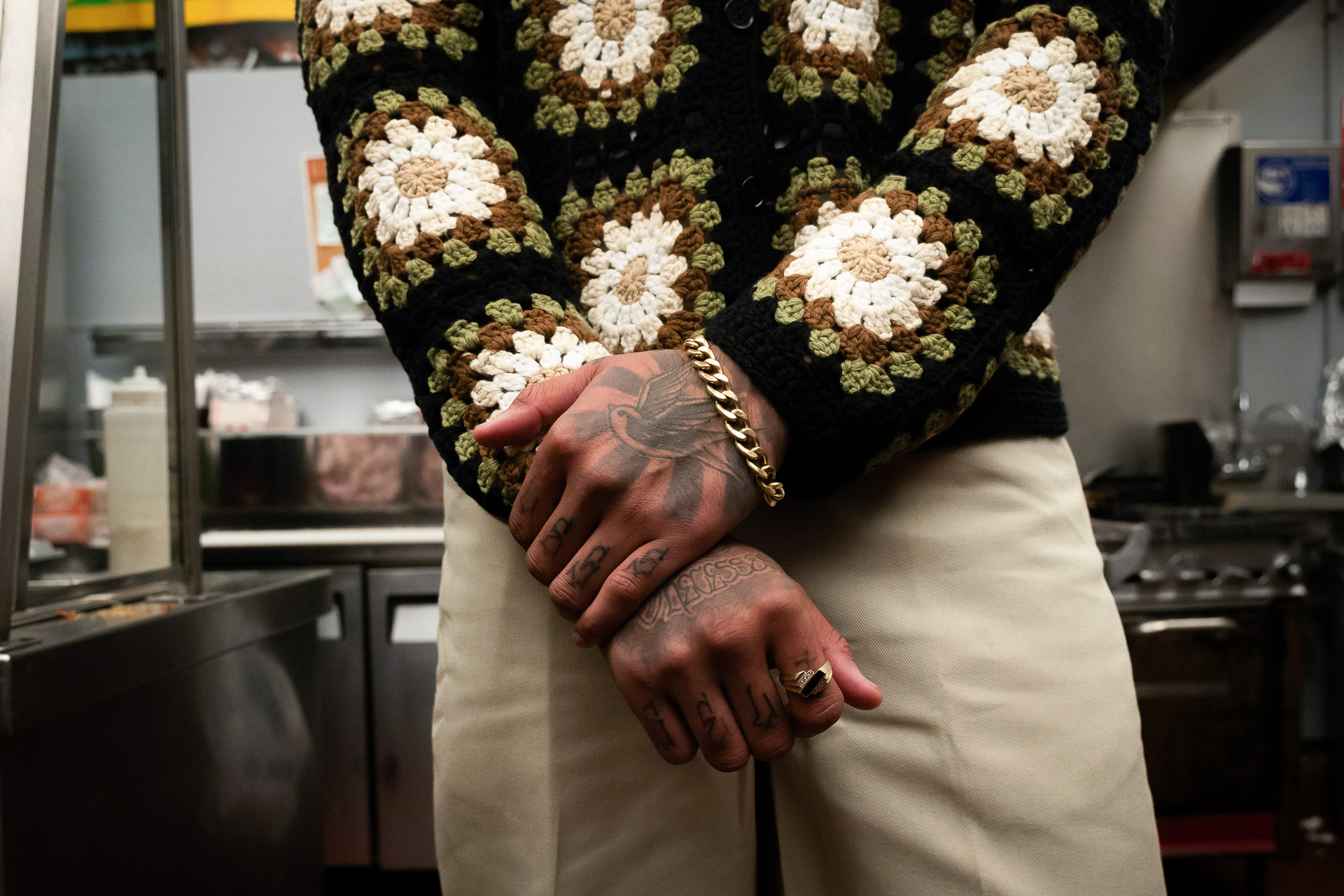 A person crosses their arms in front of their body with one hand holding their wrist. A gold bracelet hands from their wrist and tattoos cover their hands. They are wearing a knit shirt and off white pants.
