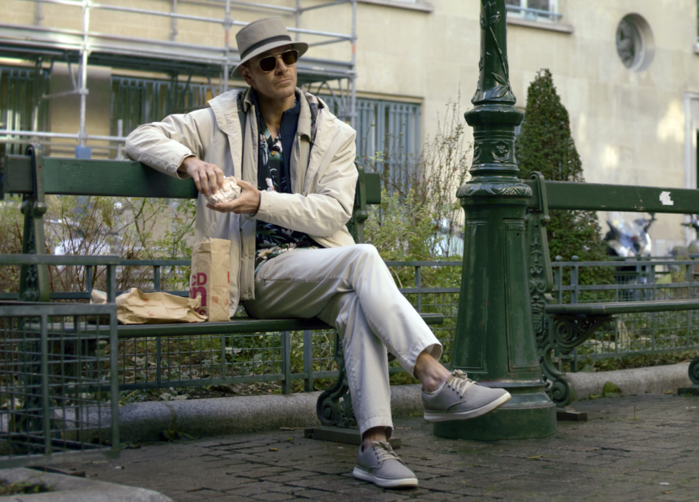A white man wearing leisure wear including white pants and a beige sun hat sits on a bench, looking relaxed.