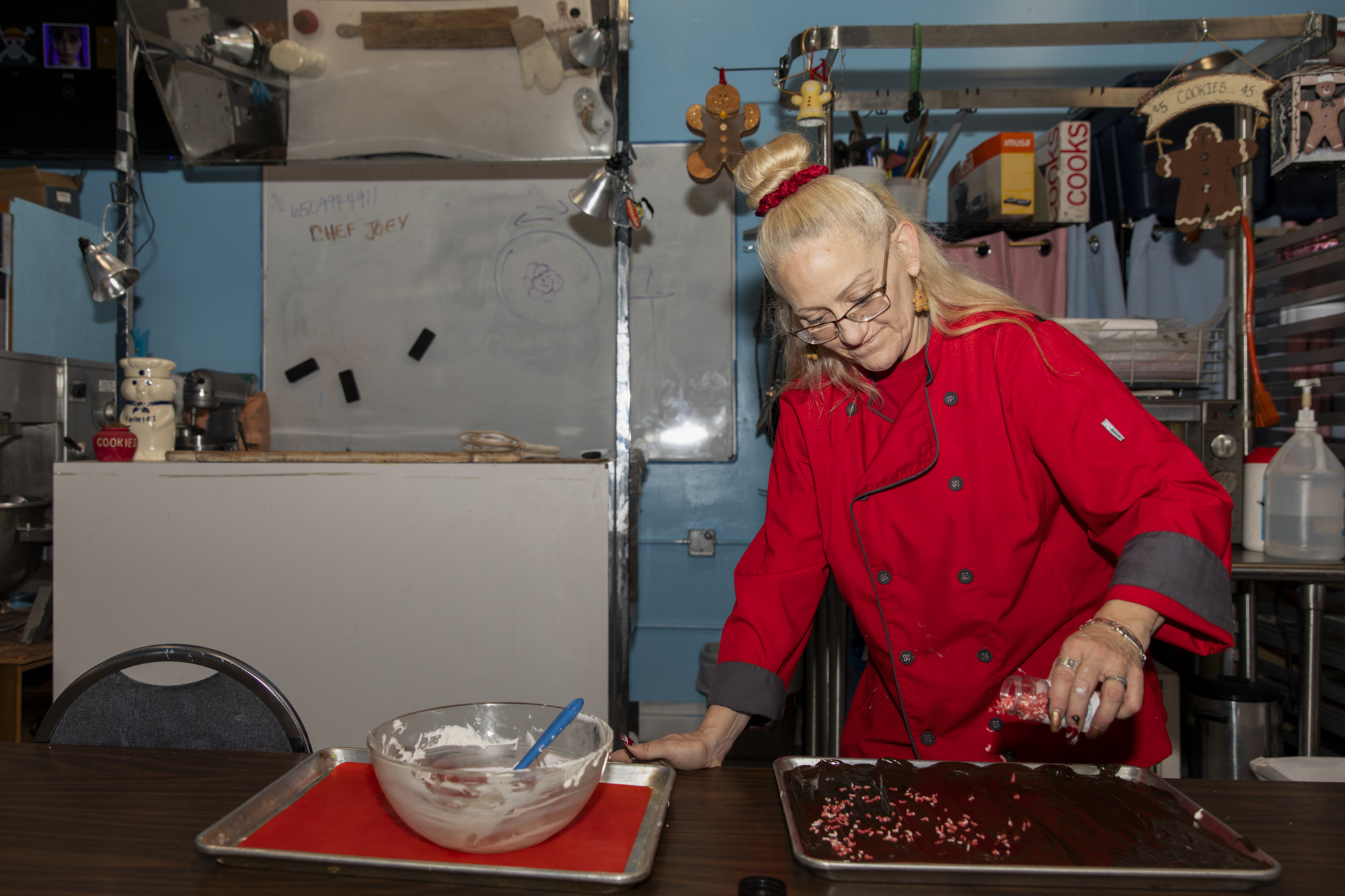 A woman in a red chef's jacket pours sprinkles over a tray of peppermint bark.