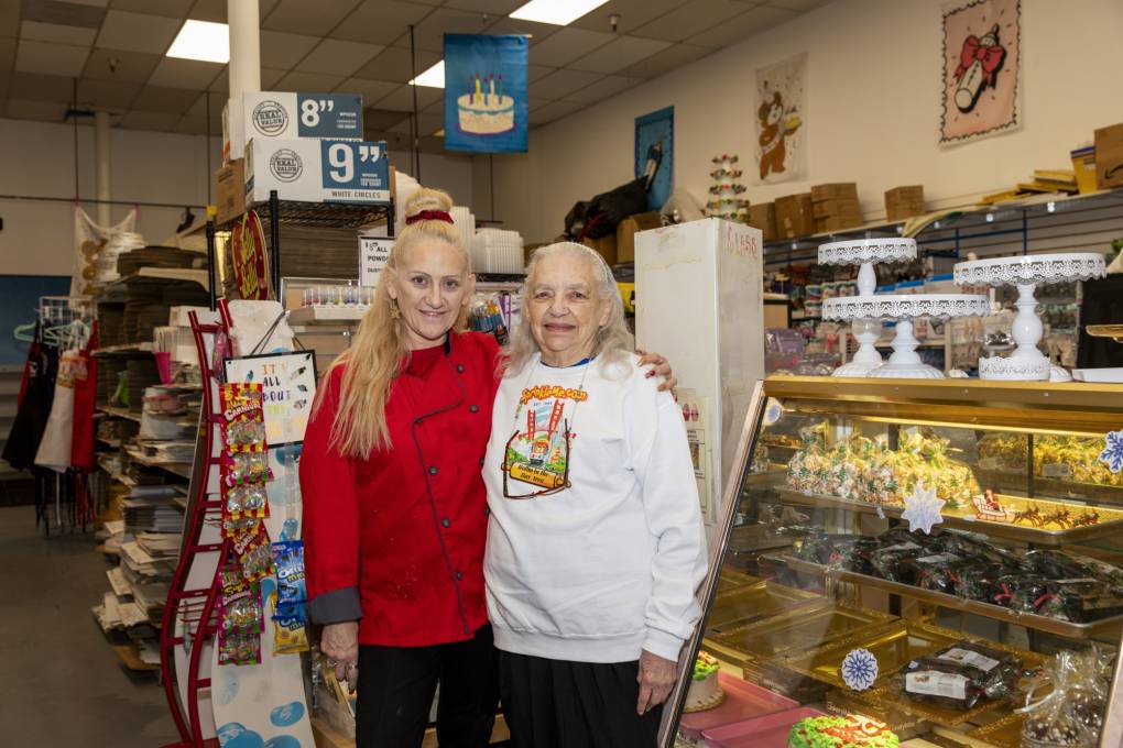 A woman in a red chef's coat poses her arm around the should of an older woman in a white sweatshirt that reads, "SprinkleMe.com." They are standing next to the front display case of a baking supply shop.