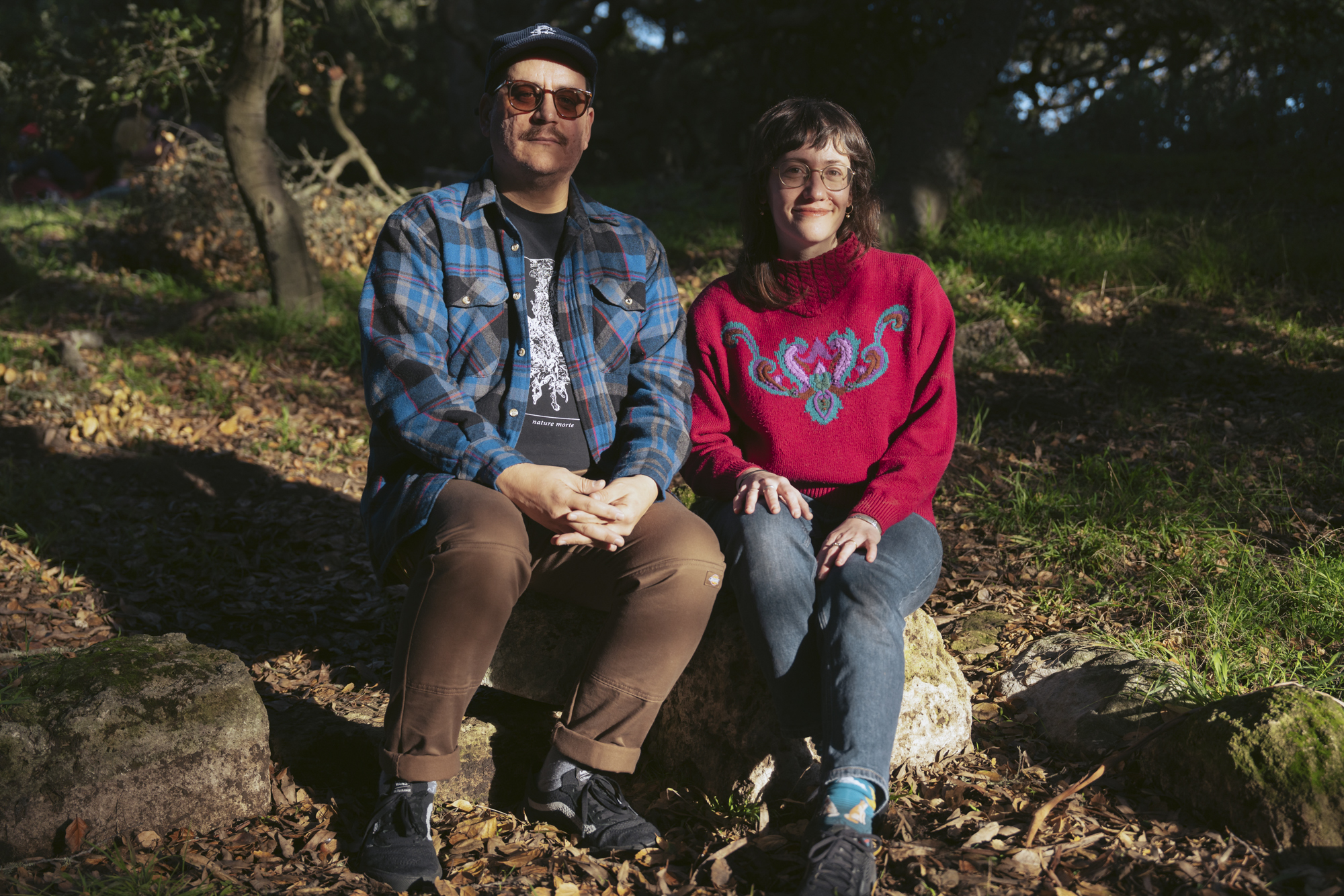 Two people sit next to each other in a wooded area, looking at the camera.
