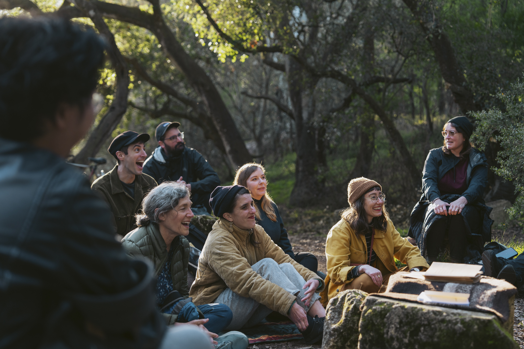 A group of people sit together smiling in a wooded area.