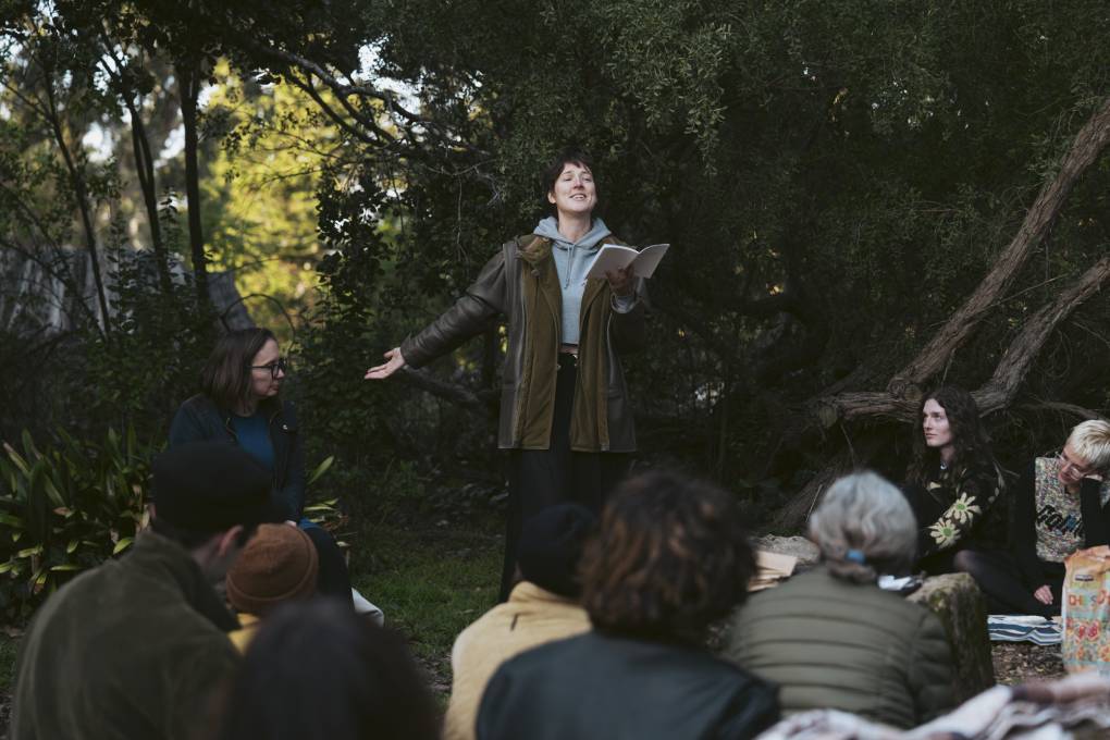A person holding a book speaks in front of a group of people in a wooded area.