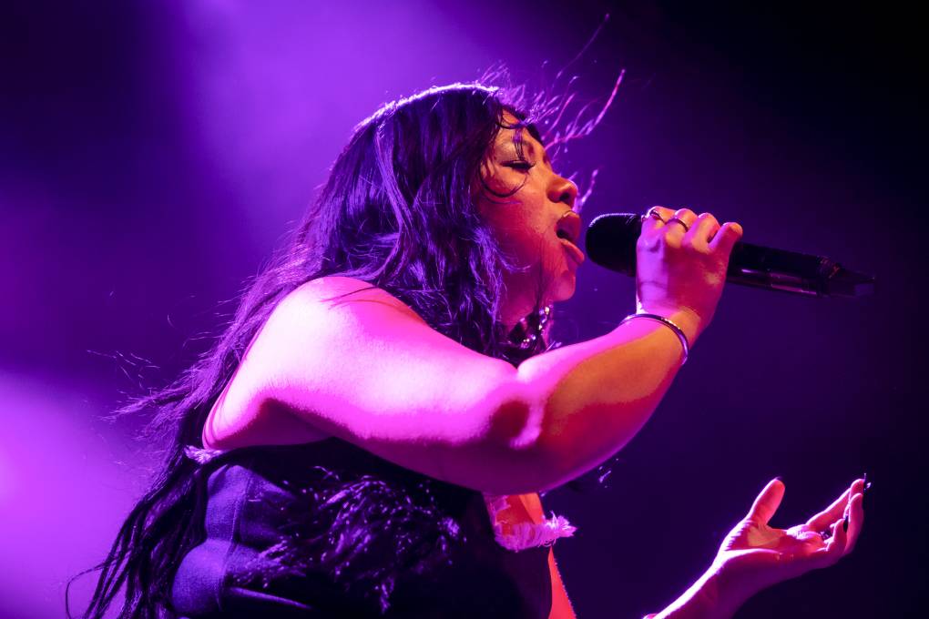 a young woman with black hair sings into a microphone bathed in purple light on stage