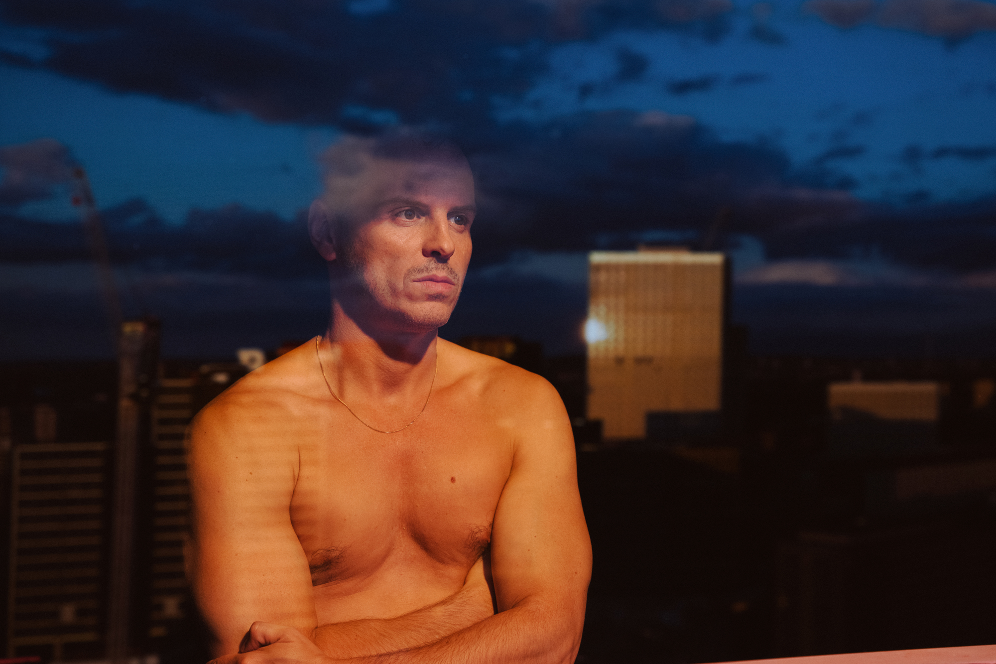 Bare-chested man lit orange looks out high rise window at reflected city