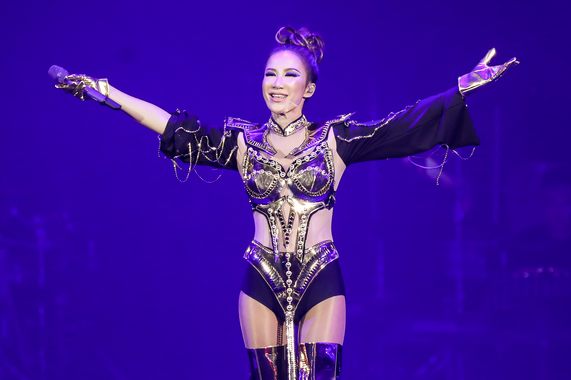 Woman in elaborate metal and black costume holds arms up, mic in one hand