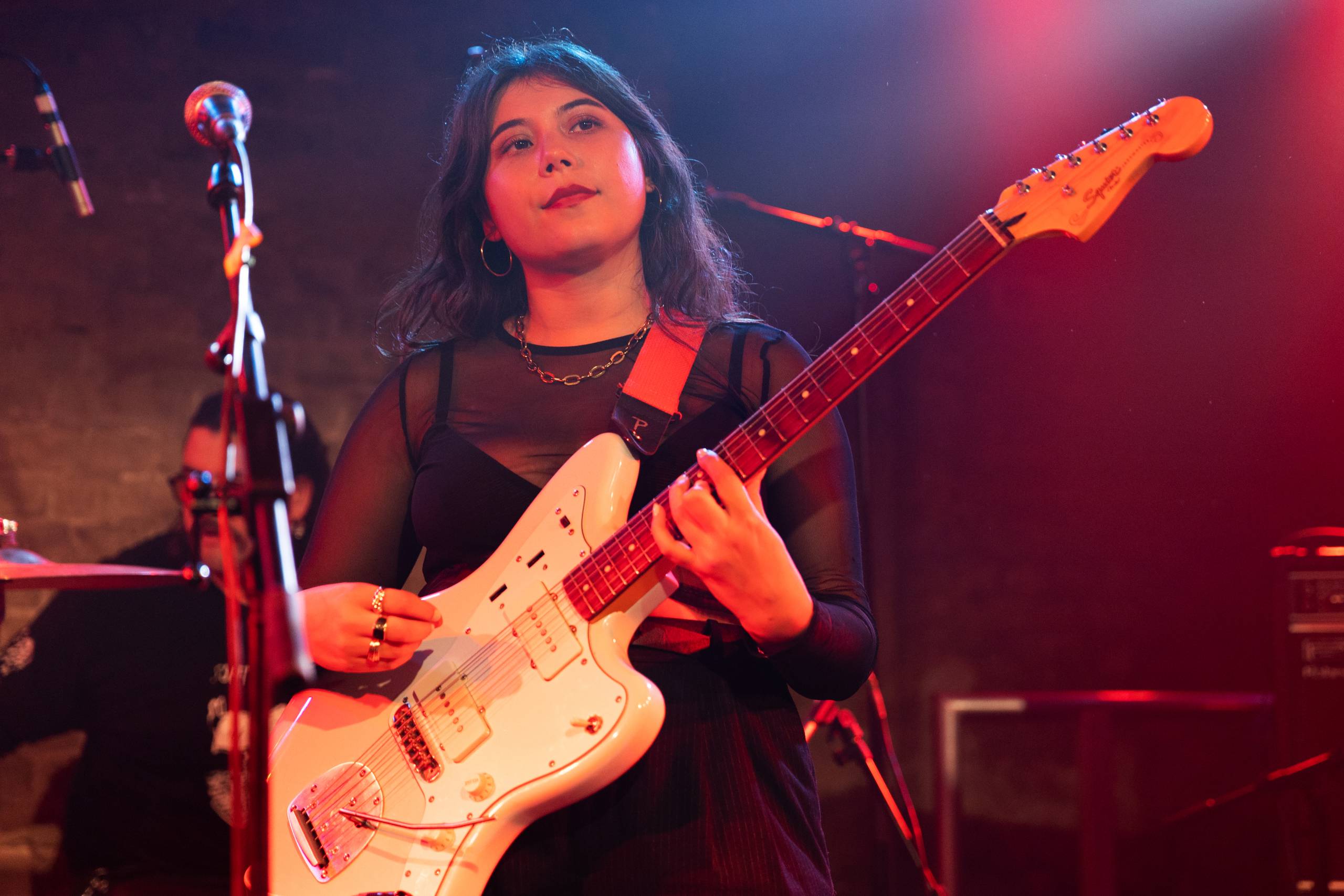 a young woman with dark hair and a dark dress plays a white guitar on stage in purple lighting