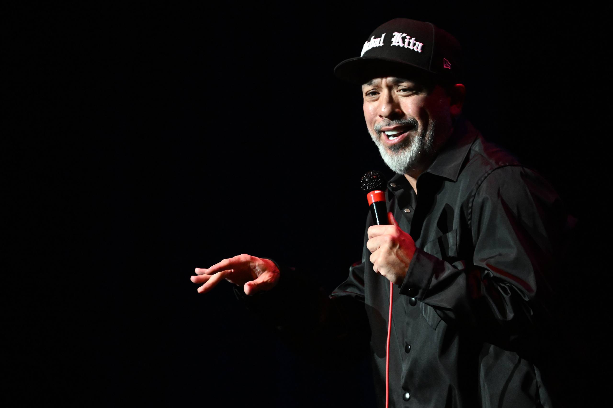 A man wearing all black and a baseball cap talks into a microphone onstage in a dark room.