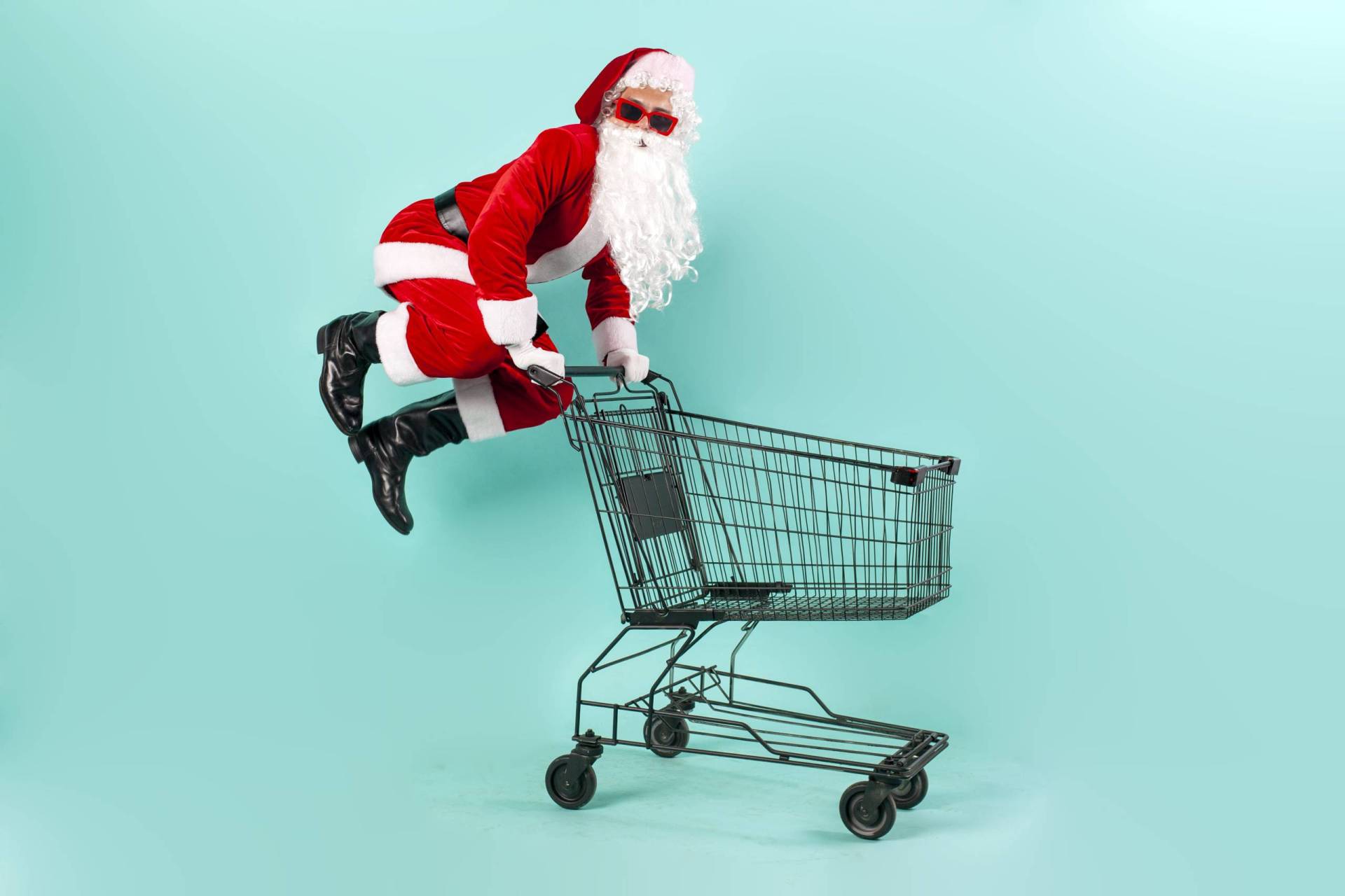 Santa Claus in sunglasses jumps in the air behind an empty shopping cart.