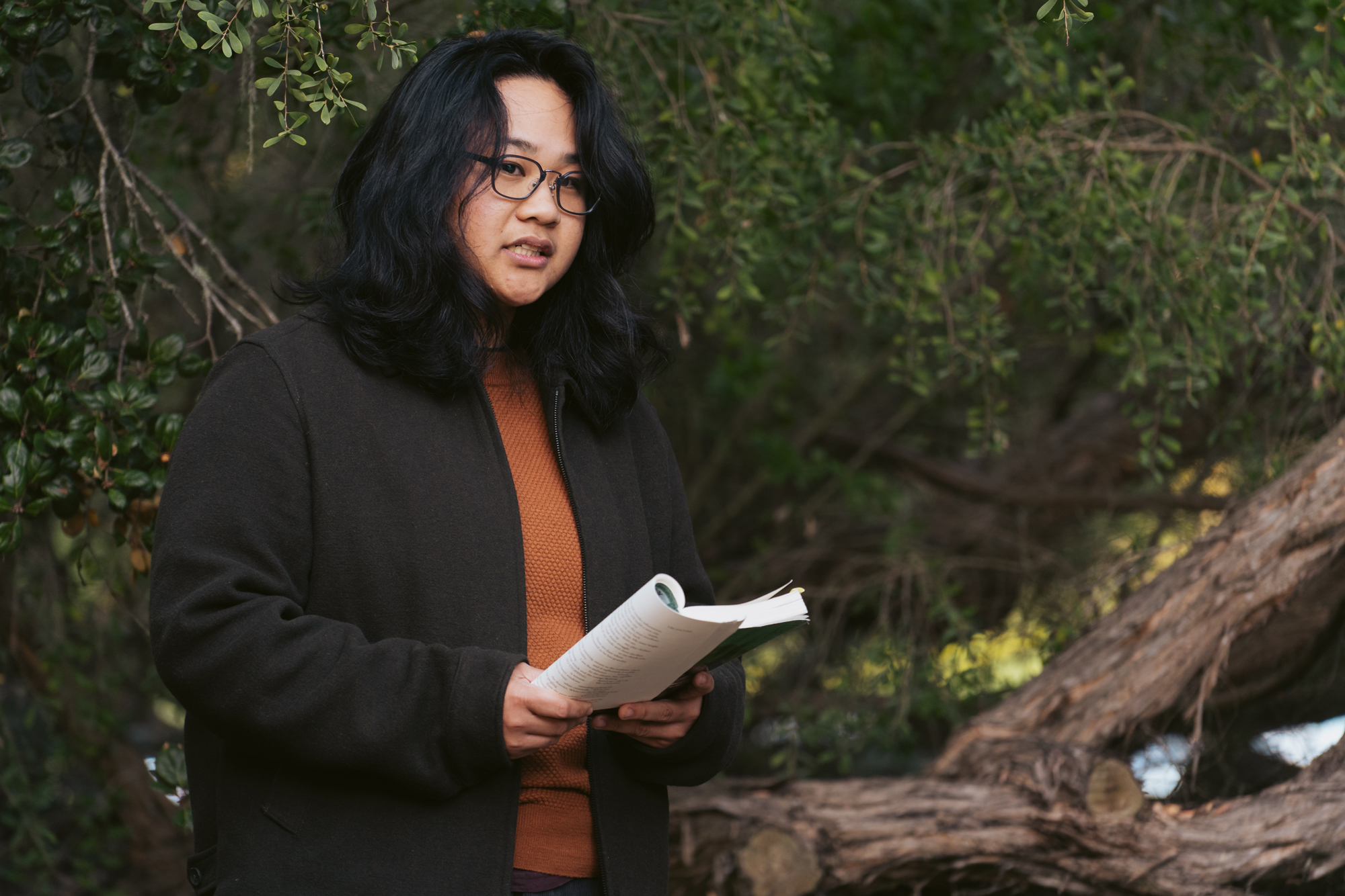 Person with shoulder-length black hair and glasses reads from paperback in wooded area