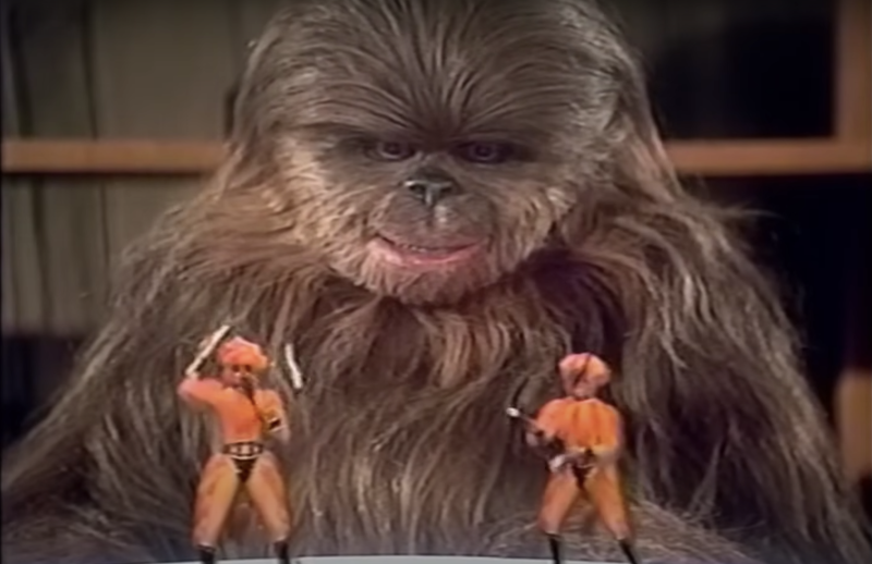 A fur covered creature sits at a desk and watches two tiny figures in orange costumes dance.