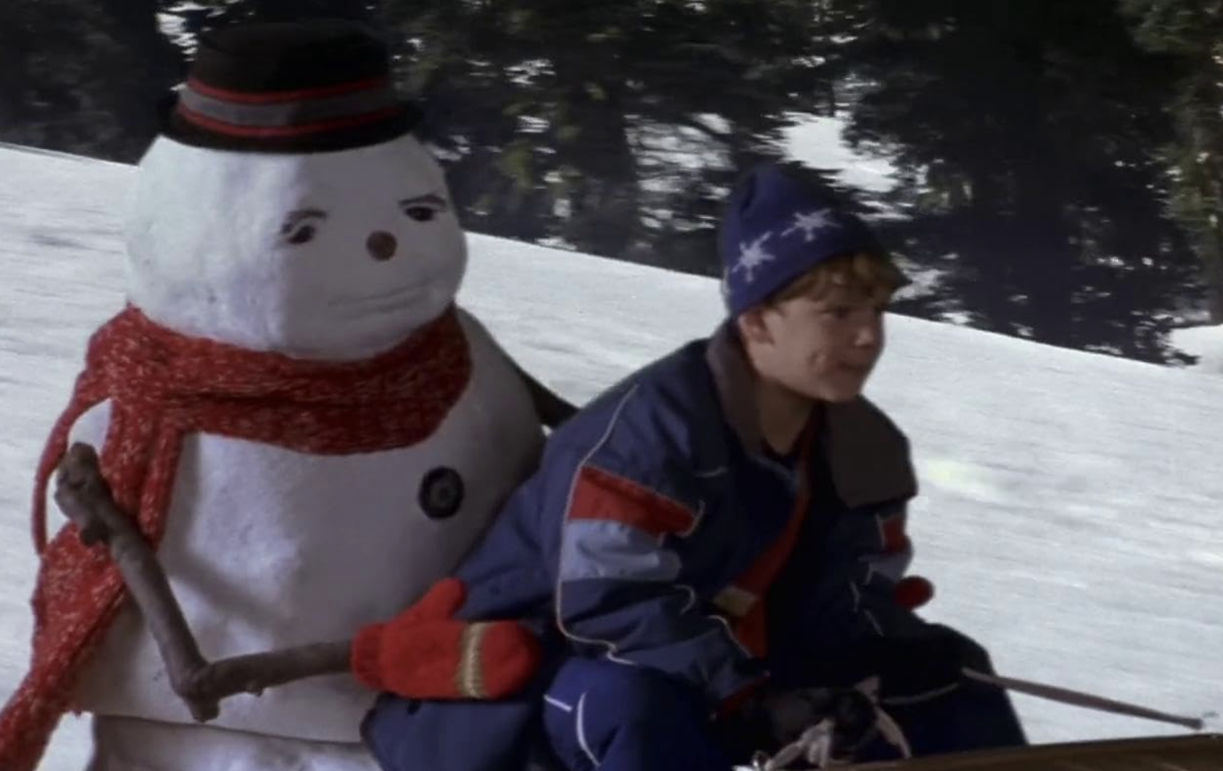 A boy in winter clothes sits on the front of a sled as it zips down a snow-covered hill. Behind him sits a snowman holding onto his waist.