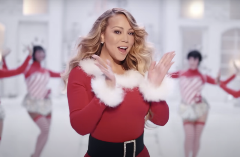 Mariah Carey in a festive red dress with white fluffy trim gesturing and singing. Dancers are behind her, out of focus.
