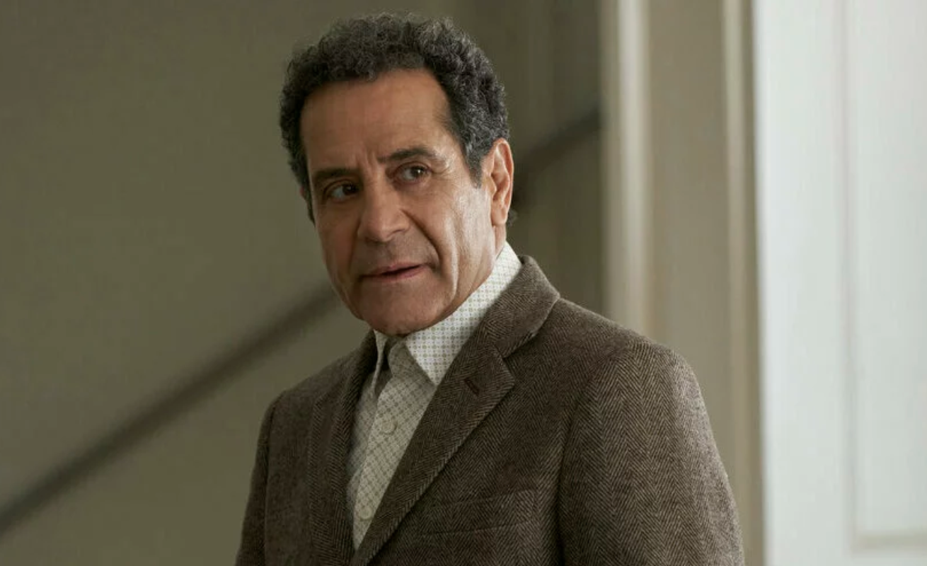 An older man with brown curly hair stands alone in a stairwell. He is wearing a brown suit jacket and white shirt with no tie.