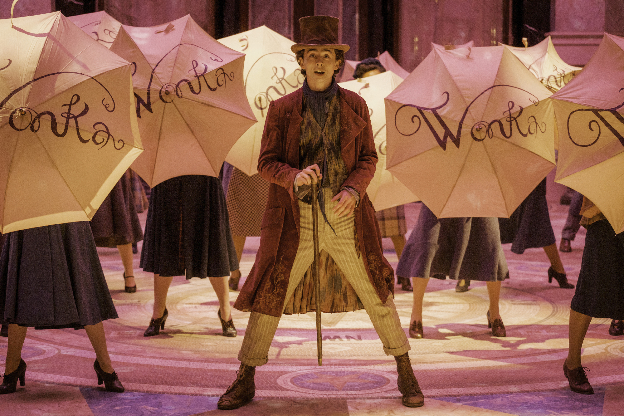 Man in top hat and Victorian attire stands with legs apart, mouth open with background dancers holding umbrellas with "Wonka" on them