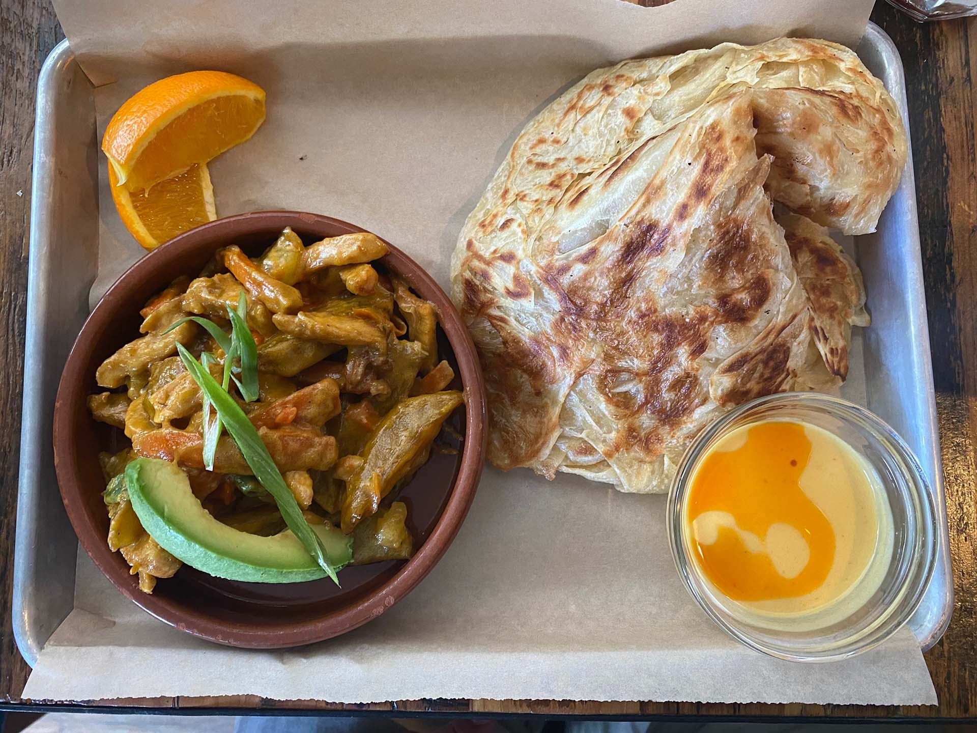 Crispy, flaky roti (Malaysian flatbread) served alongside a bowl of curried chicken and root vegetables on a paper-lined metal tray.