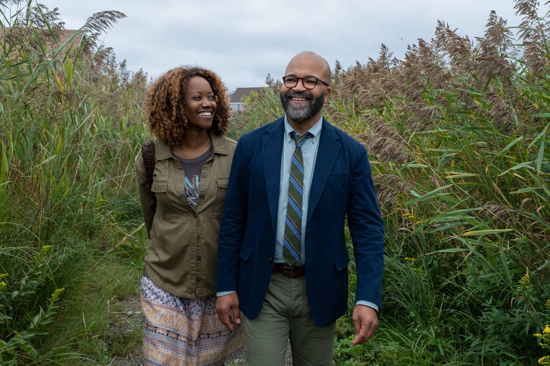 A Black woman and man walk down a grassy path, surrounded on both sides by tall shrubs and plants. They are smiling.