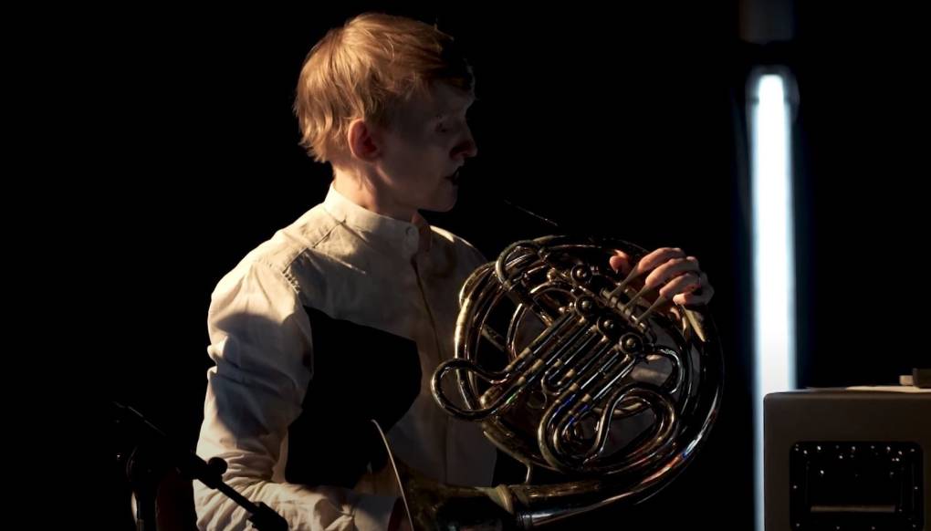 Andy Guthrie plays a French horn on a dimly lit stage.