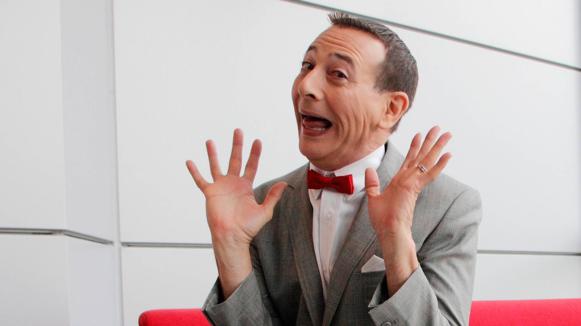 A man in a grey suit and red bowtie gestures wildly, mouth agape.