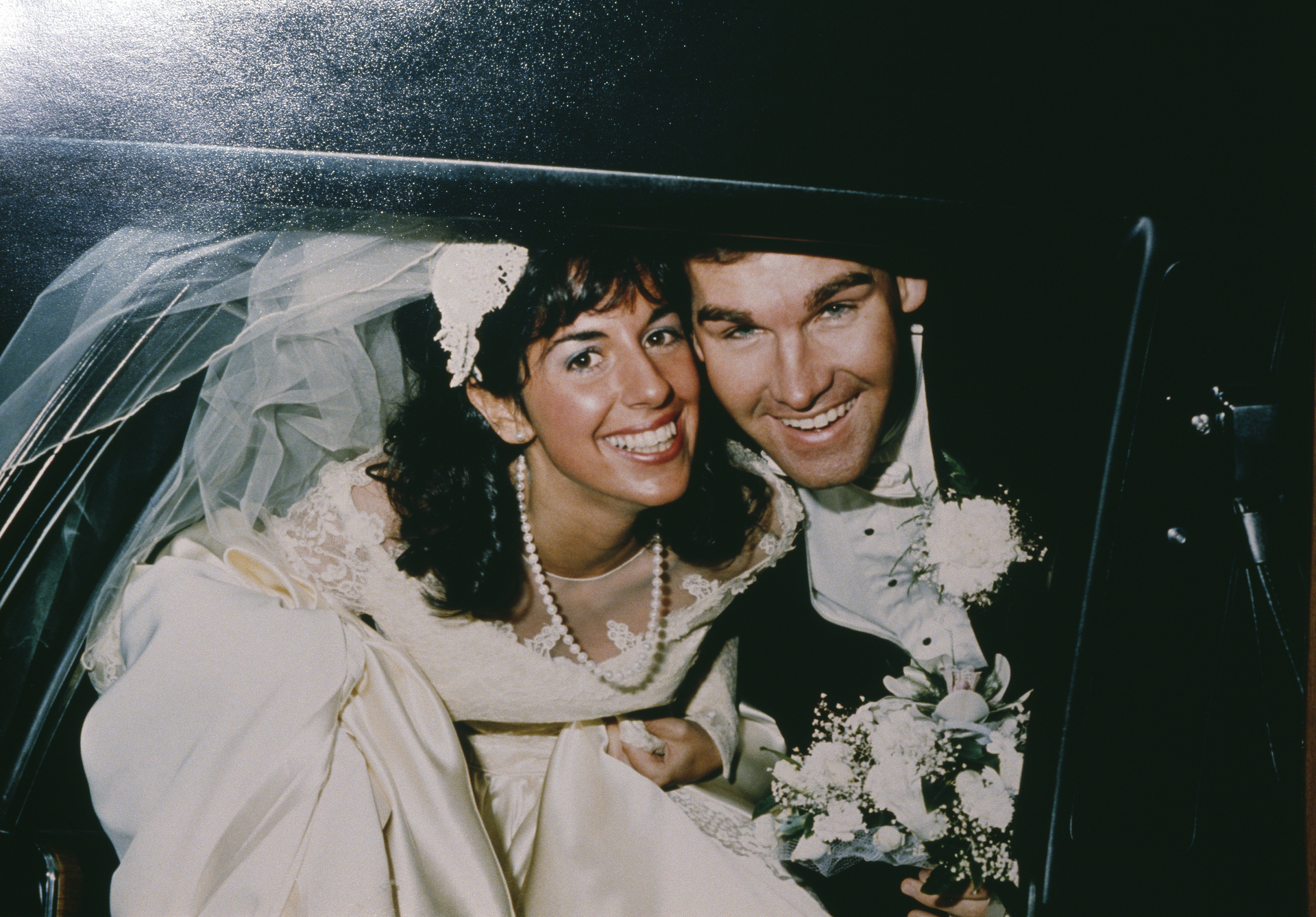 A bride and groom smile broadly from the back of a car.
