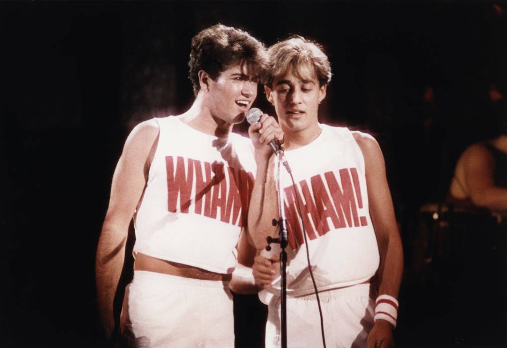 Two young men stand close together behind a microphone. They are wearing white pants and white cropped tops that say WHAM! on them.