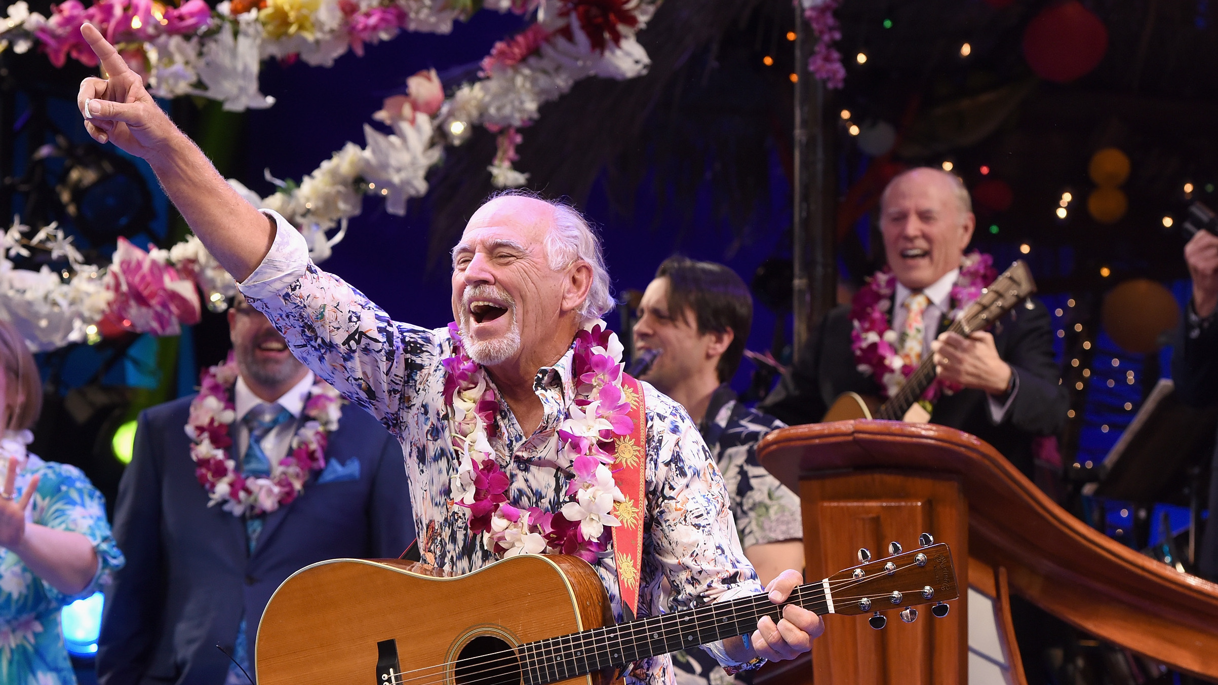Man in hawaiian shirt and flower lei holds guitar in one hand and points finger up, mouth open