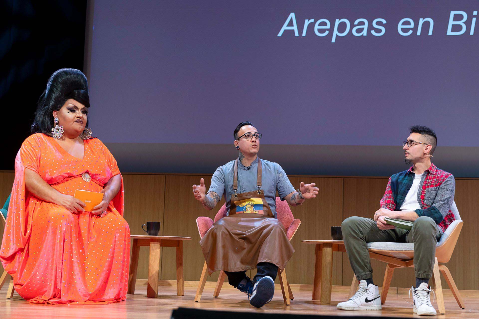 A drag performer in a bright orange dress, a chef wearing glasses, and a journalist in white sneakers sit on stage. The words "Arepas en Bici" are projected onto the screen behind them.