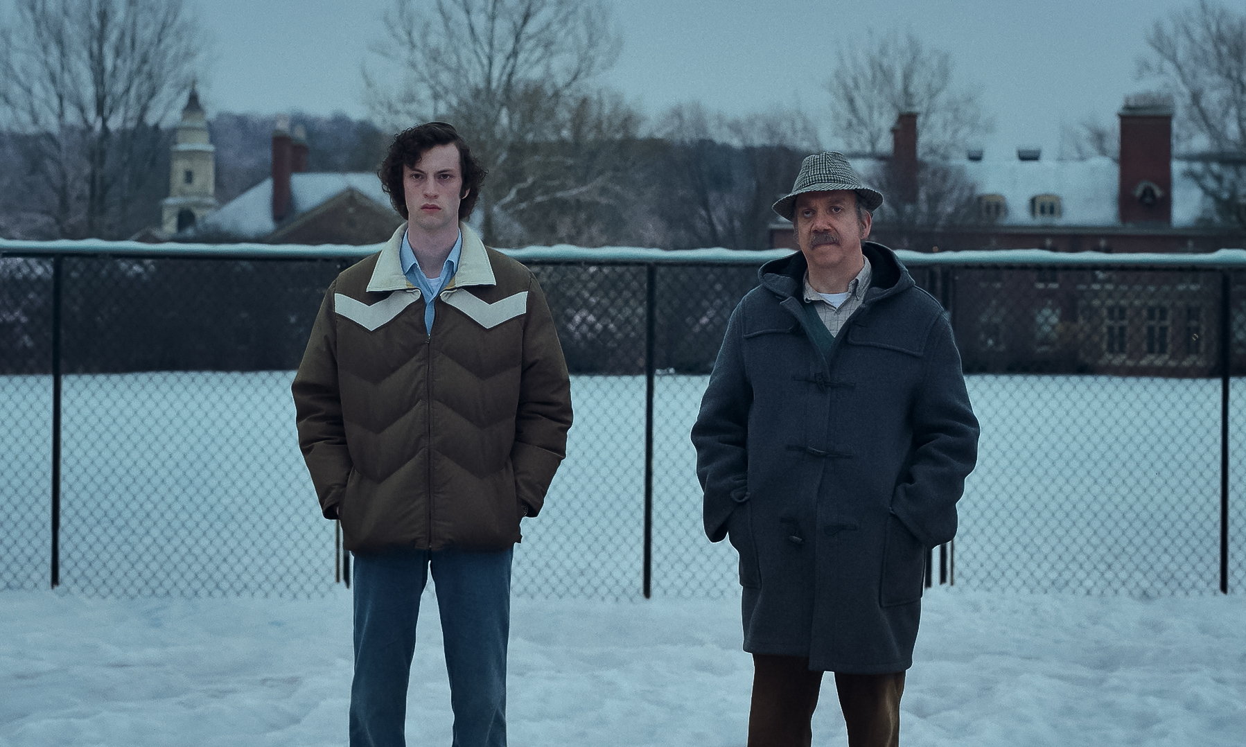 A tall young man and a shorter, older man with a mustache stand side by side in a snowy field. 