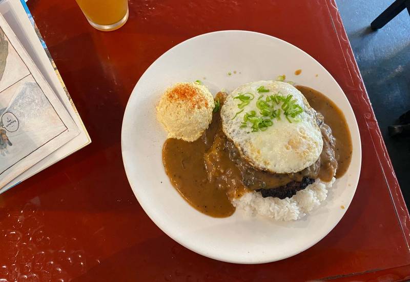 A plate of gravy-drenched Hawaiian loco moco with two burger patties, a runny egg, a big mound of white rice and a scoop of potato salad.