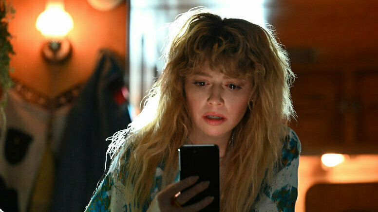 A woman with long shaggy blond hair stares worried into a phone screen. 