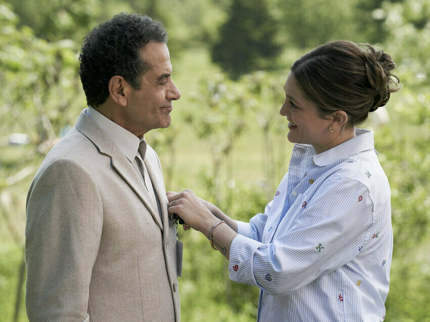 A smiling woman wearing a white shirt places something in the jacket pocket of an older man.
