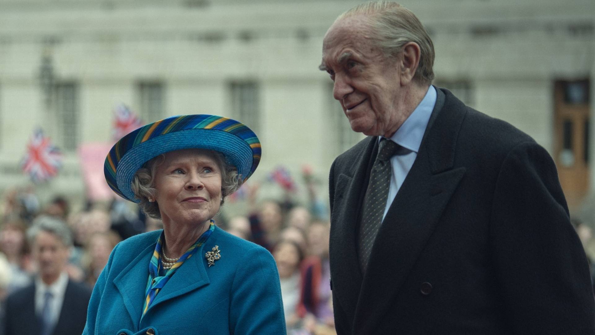 An elderly woman in a blue suit and matching hat looks up fondly at an elderly man in a suit.