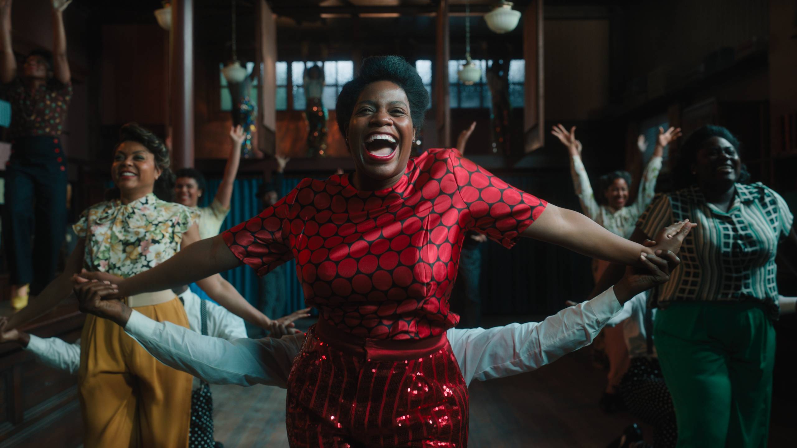 a young Black woman in a red dress extends her arms while singing, while two other Black women sing and dance in the background
